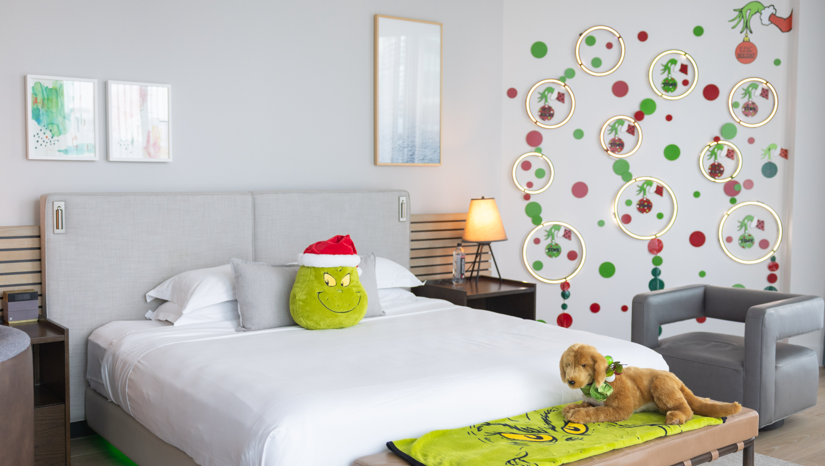 Grinch-themed bedroom with a bed, Grinch pillow, dog plush on a blanket, and a wall decorated with Grinch hands and lights