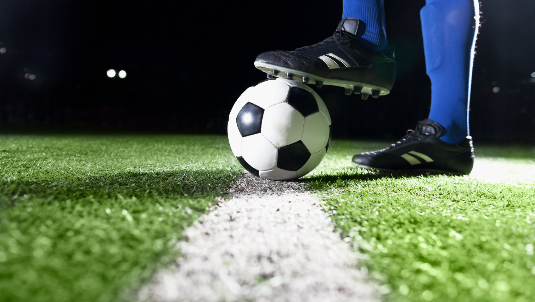 Close-up of a soccer player's foot on a ball on a vibrant green turf field with a white line at night